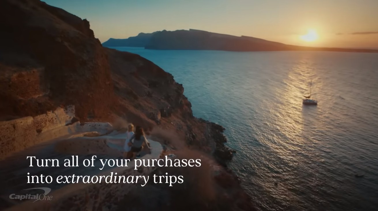 A still from a Capital One Venture X advertisement. Two people walk down a cliffside path above the sea at sunset, a sailboat anchored below, under the line "Turn all of your purchases into extraordinary trips"